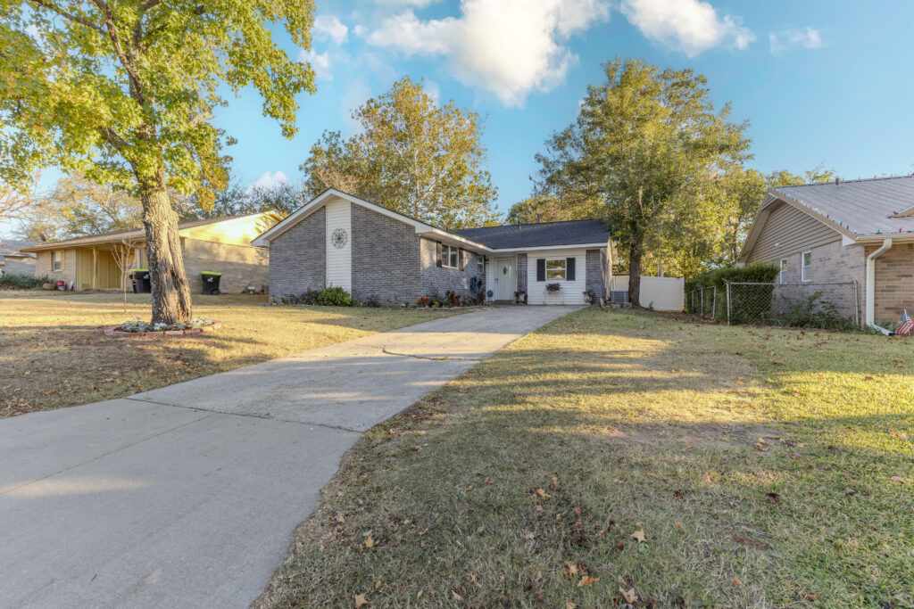 The front of 917 W 24th Street in Ada, OK features a classic single-story design with gray brick and white siding, a spacious driveway, and a well-kept yard shaded by mature trees — giving this home a welcoming, peaceful curb appeal.