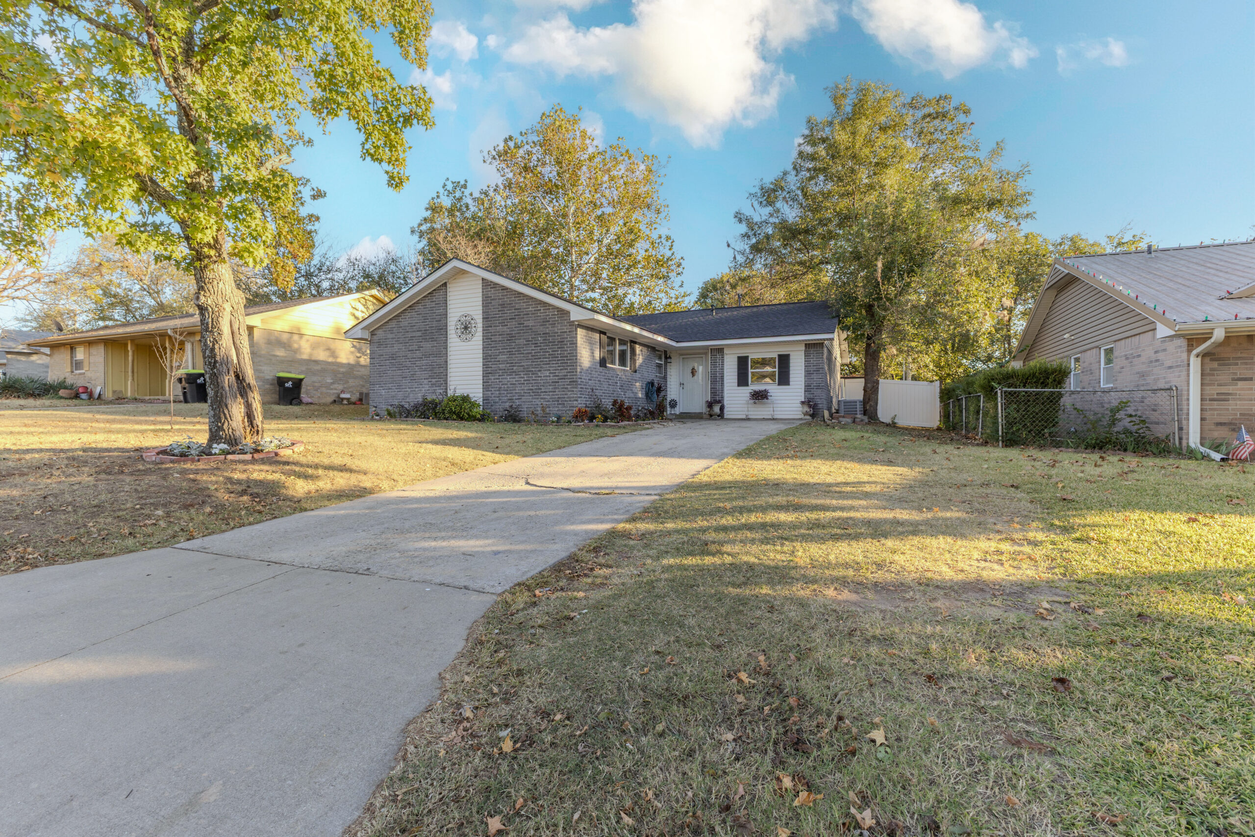 The front of 917 W 24th Street in Ada, OK features a classic single-story design with gray brick and white siding, a spacious driveway, and a well-kept yard shaded by mature trees — giving this home a welcoming, peaceful curb appeal.
